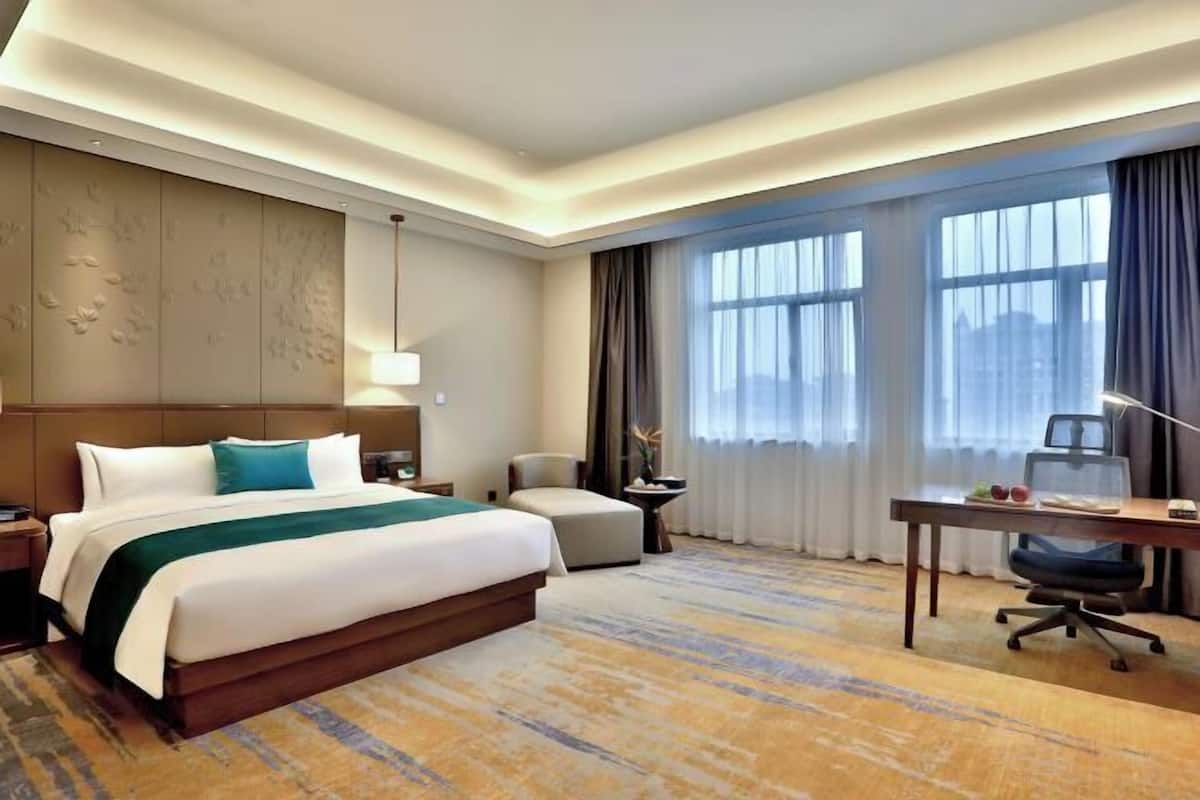 A hotel room with a large bed, a desk, a chair, a nightstand, a lamp, and a view of the city through large windows.