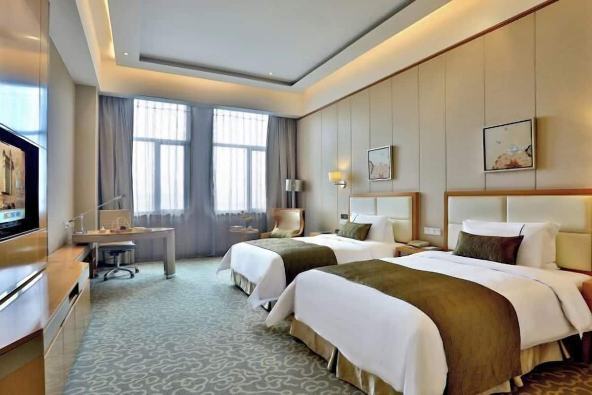 A hotel room with two beds, a desk, a television, and a large window with curtains.