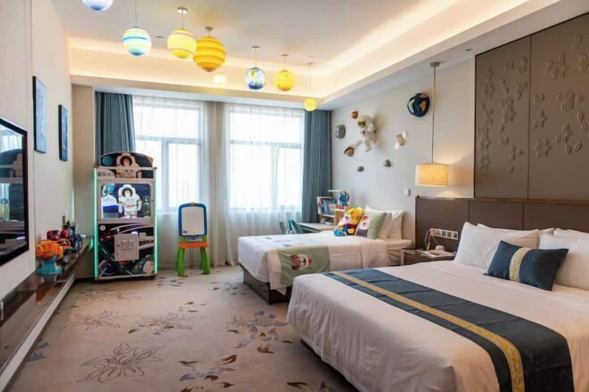 A hotel room with two beds, a desk, a chair, a TV, and a bookshelf.