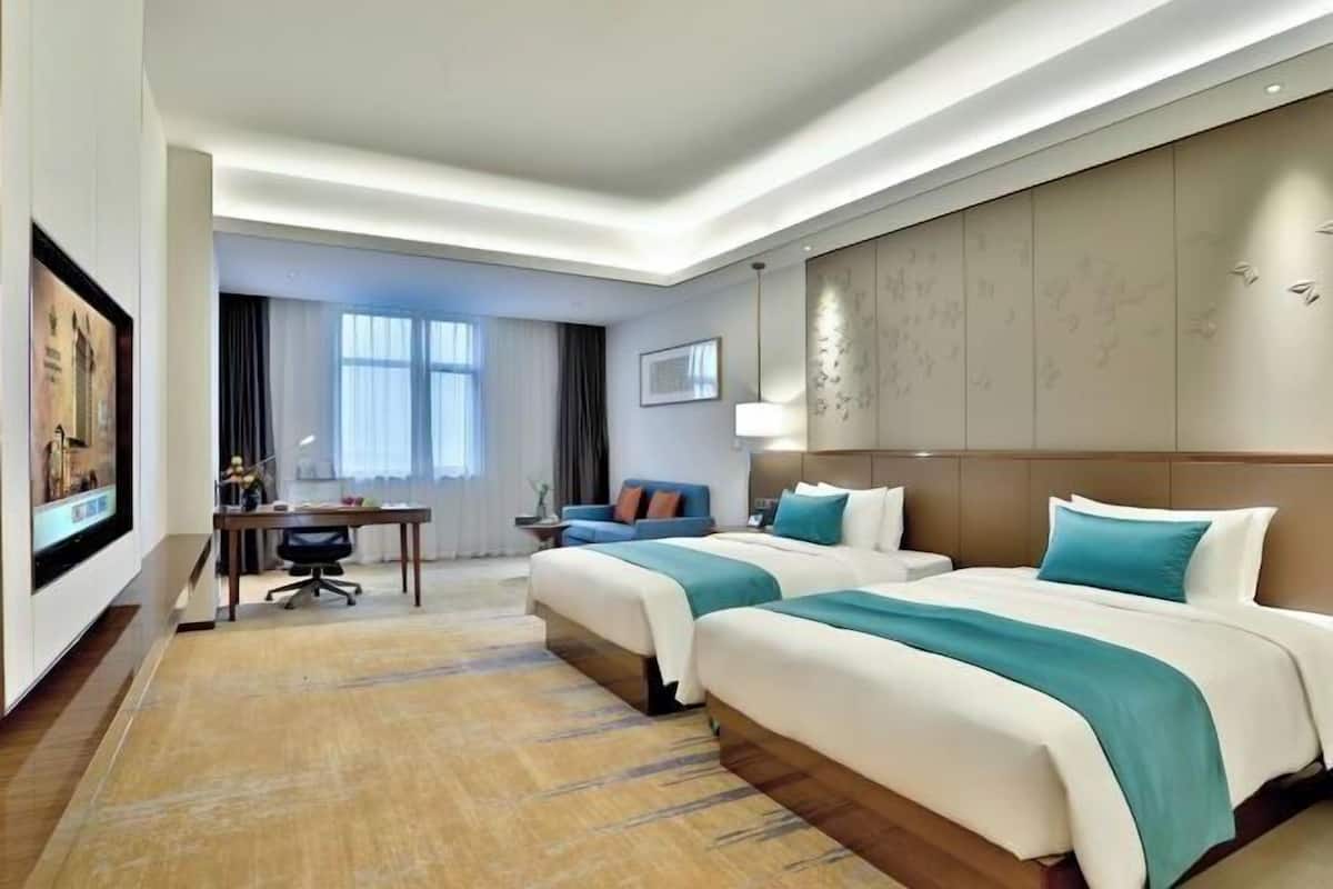 A hotel room with two beds, a desk, a chair, a large window, and a wall-mounted artwork.