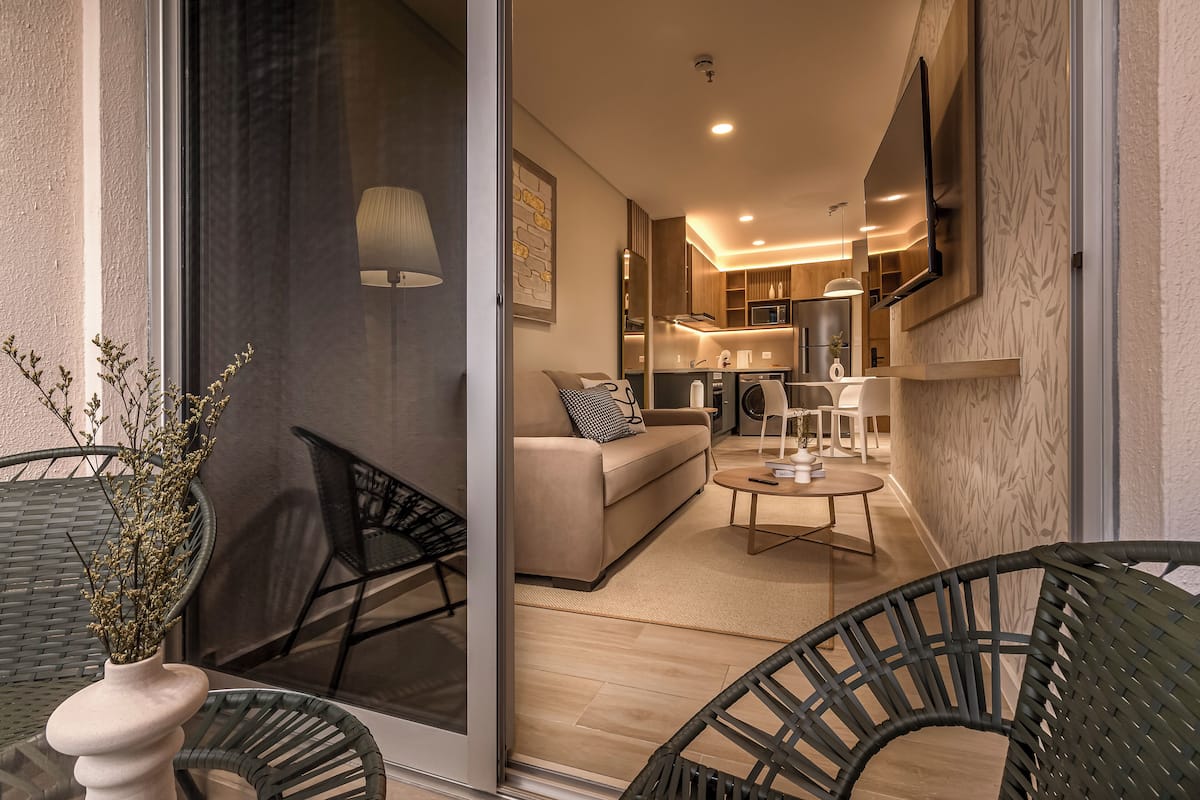 A modern living room with a sofa, coffee table, and chairs, leading to a kitchen area with a TV and modern appliances.