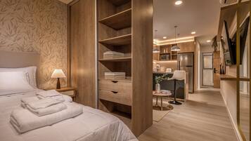 A modern bedroom with a large bed, wooden wardrobe, and a small kitchenette in the background.