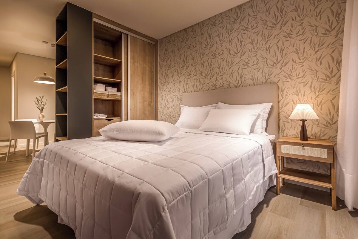 A neatly made bed with a headboard, a bedside table with a lamp, and a wardrobe with shelves.