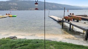 Muelles que se adentran en un lago tranquilo con un farol y un mástil, rodeados de zonas verdes y una colina boscosa al fondo.
