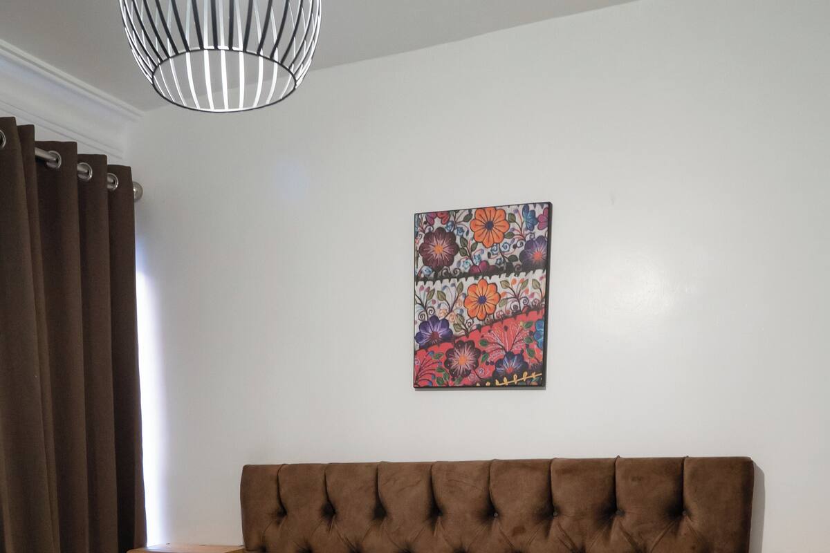 Una cama bien hecha con una colcha estampada, dos almohadones decorativos y una obra de arte colgada en la pared.