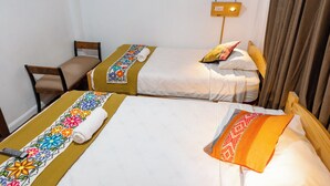 Habitación de hotel con dos camas individuales, cada una con una colcha de colores y una toalla enrollada. Hay una silla de madera y una mesita de noche con una lámpara.