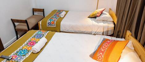 A hotel room with two single beds, each with a colorful bedspread and a rolled towel. There is a wooden chair and a bedside table with a lamp.