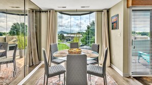 Un comedor moderno con una mesa y sillas de vidrio, vista a un jardín y montañas, y un gran ventanal corredizo al exterior.