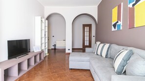 A modern living room with a grey sofa, a TV, a white shelving unit, and a wooden floor.