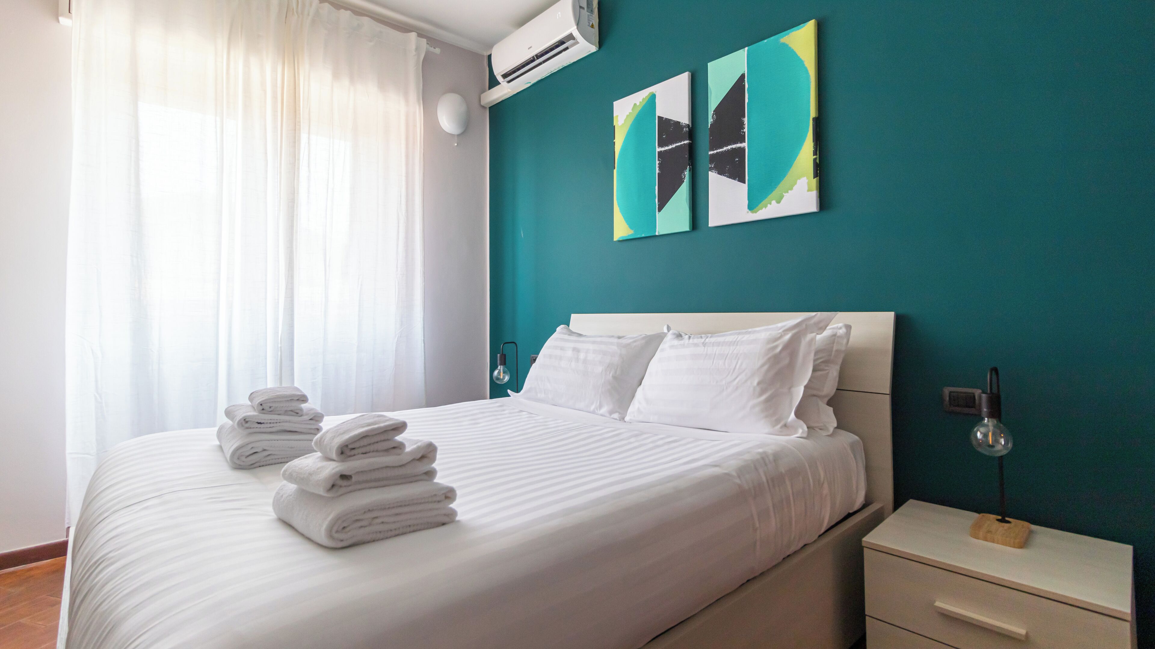 Een netjes opgemaakt bed met opgevouwen handdoeken, een nachtkastje met een lamp, en twee abstracte schilderijen aan de blauwgroene muur.