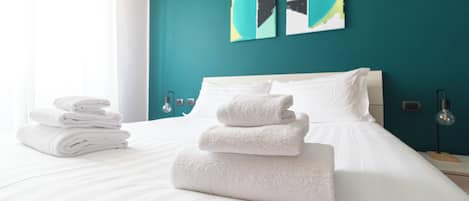 A neatly made bed with white linens and towels, a teal accent wall, and modern decor.