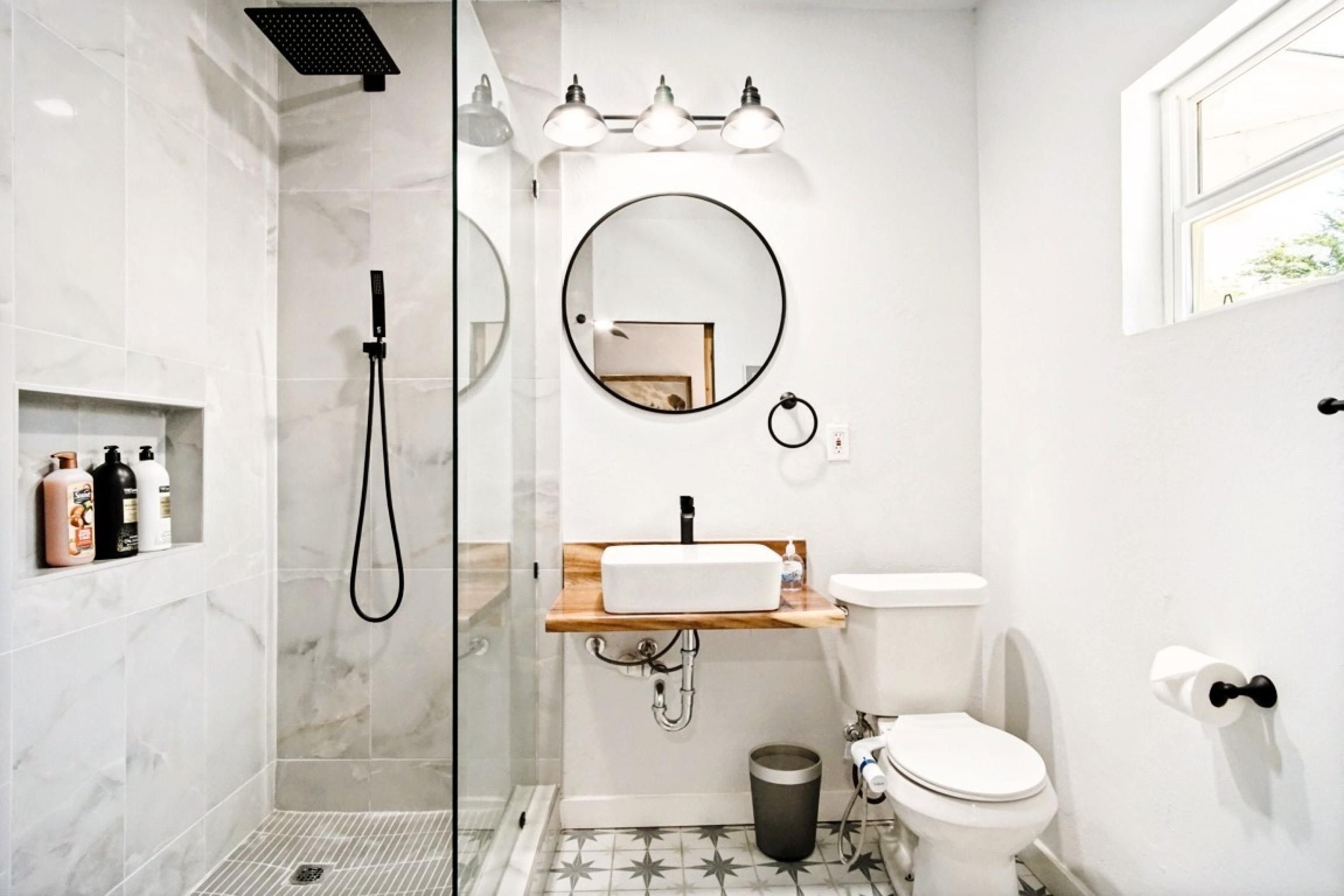 A modern bathroom with a glass-enclosed shower, a round mirror, a sink with a wooden countertop, and a toilet.