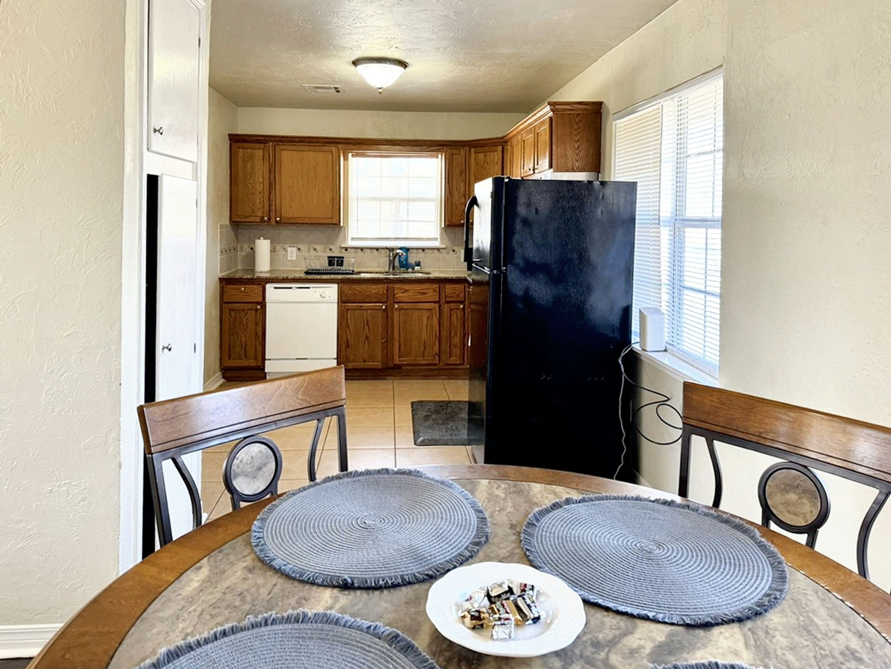 Una cocina con armarios de madera, un refrigerador negro, un lavavajillas blanco y una mesa de comedor con individuales y un plato de comida.