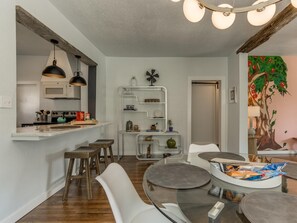 A modern kitchen with a white countertop, wooden stools, and a dining area with a glass table and chairs. - 2BR Stylish Retreat in Midtown (Oklahoma City)
