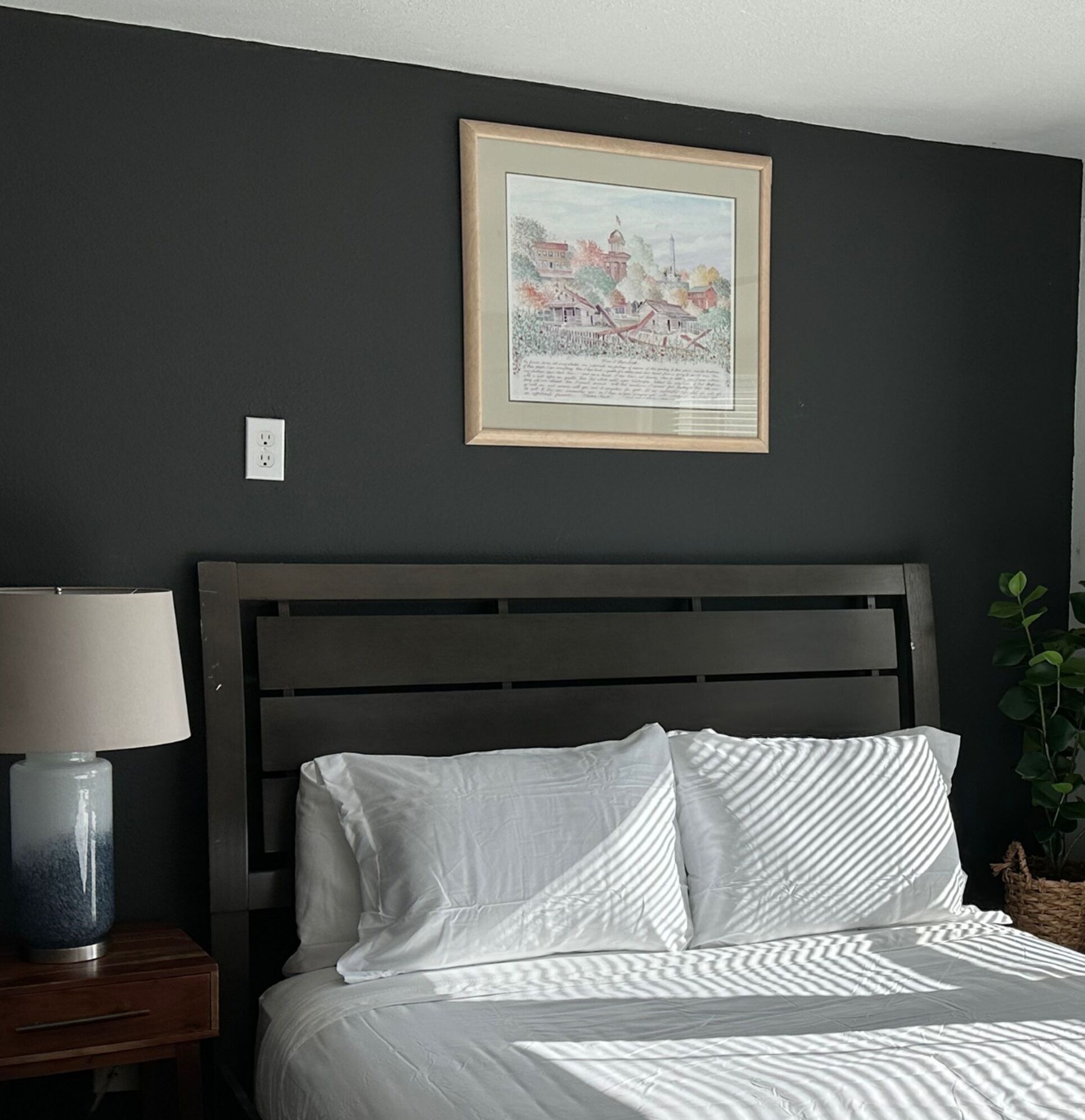 Een slaapkamer met een bed, een nachtkastje, een lamp, een lijst met een foto aan de muur en een plant.