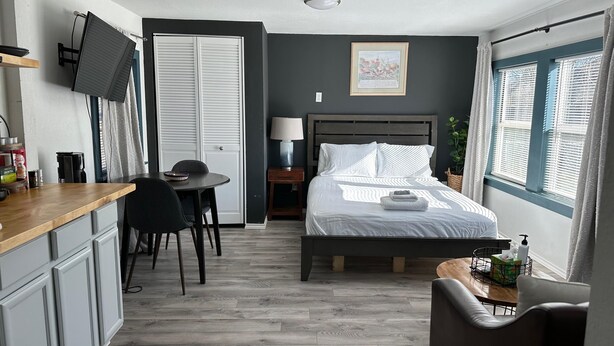 A compact hotel room with a bed, bedside table, desk, chair, and a small window with curtains.