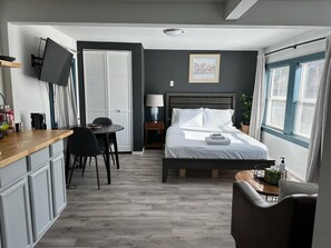 A compact hotel room with a bed, bedside table, desk, chair, and a small window with curtains. - Cute Tiny House With Fenced Yard Near Plaza Dist (Oklahoma City)