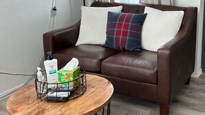 A brown leather sofa with two pillows, a black floor lamp, a framed cityscape painting, and a small wooden table with a tissue box and hand sanitizer.