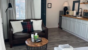 A cozy living room with a brown leather sofa, a round wooden coffee table, and a bed with neatly folded towels. A kitchen area with white cabinets and a microwave is visible in the background.