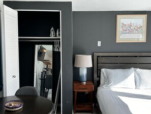 A hotel room with a bed, a bedside table with a lamp, a chair, a dining table with a basket, and a closet with a TV. - Cute Tiny House With Fenced Yard Near Plaza Dist (Oklahoma City)