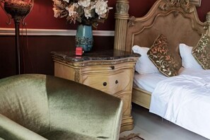 Ein Bett mit aufwendiger Kopfteinlage, ein Nachttisch mit Blumenvase und ein mit grünem Stoff bezogener Sessel.