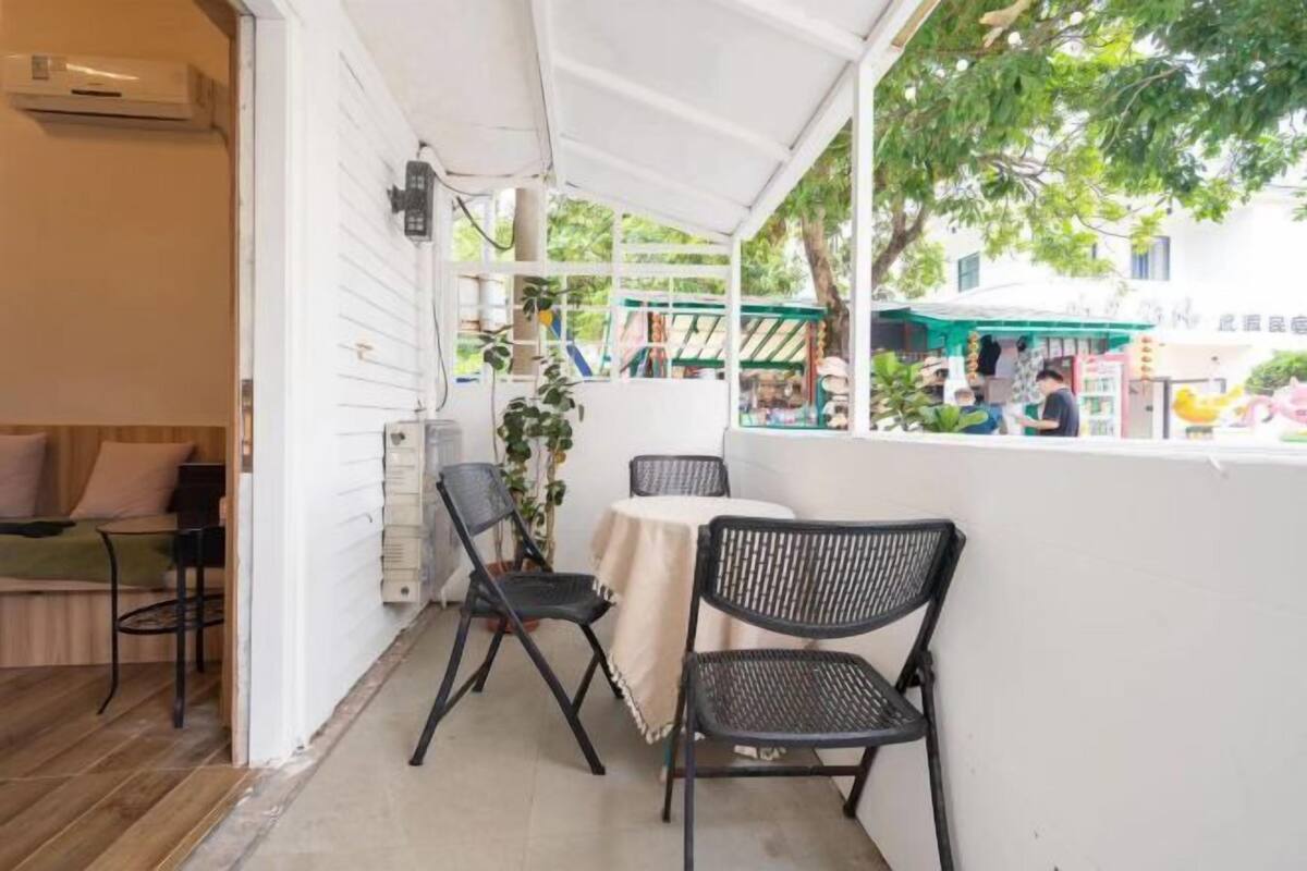 A balcony with two black chairs and a table, white walls, and a view of a street with shops and people.