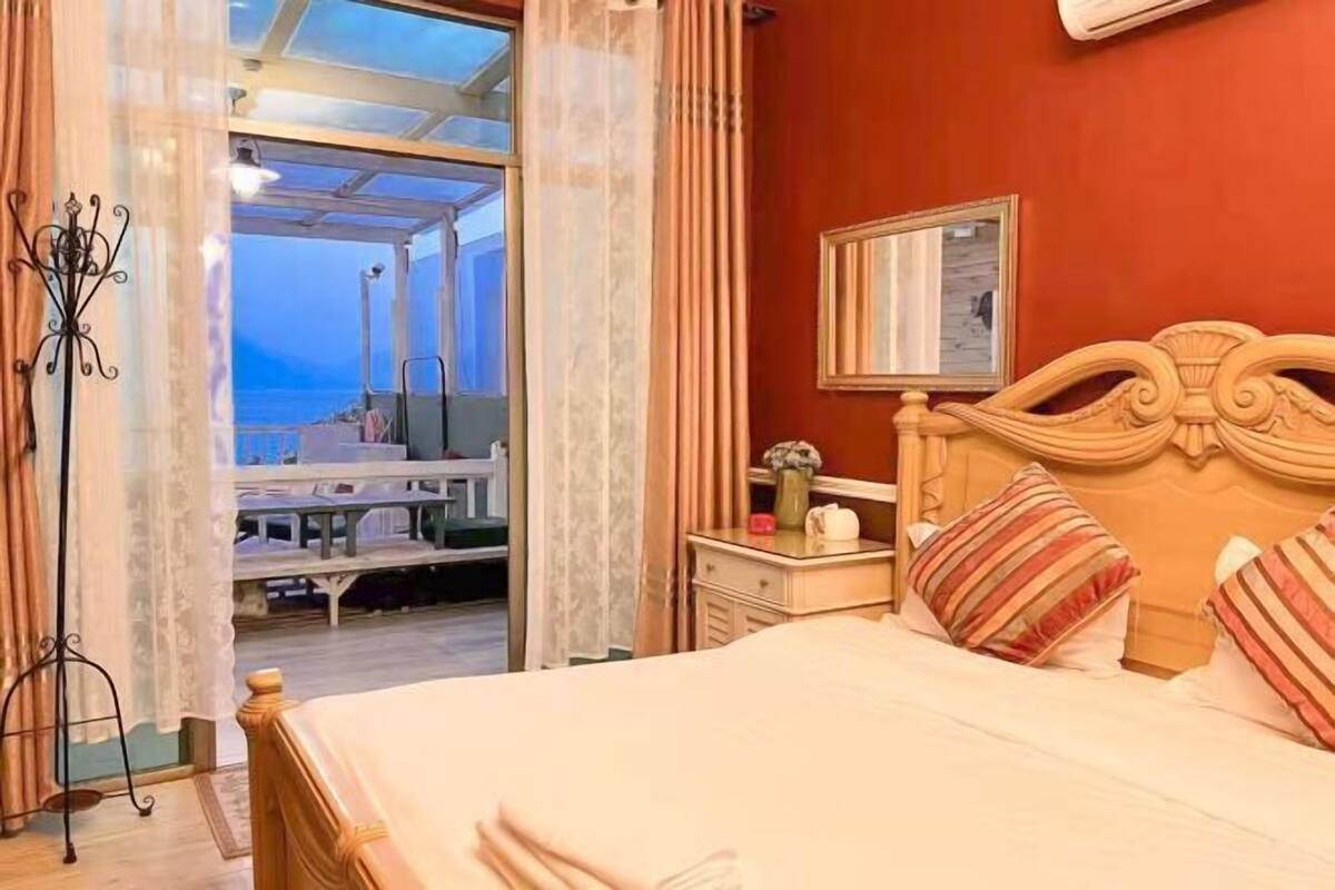 A bedroom with a bed, a nightstand, a mirror, and a view of the sea through an open door.