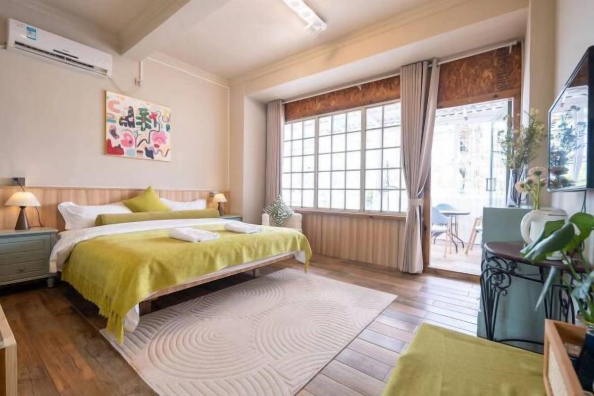 A hotel room with a bed, bedside tables, a window with curtains, a small table with a plant, and a wall-mounted air conditioner.