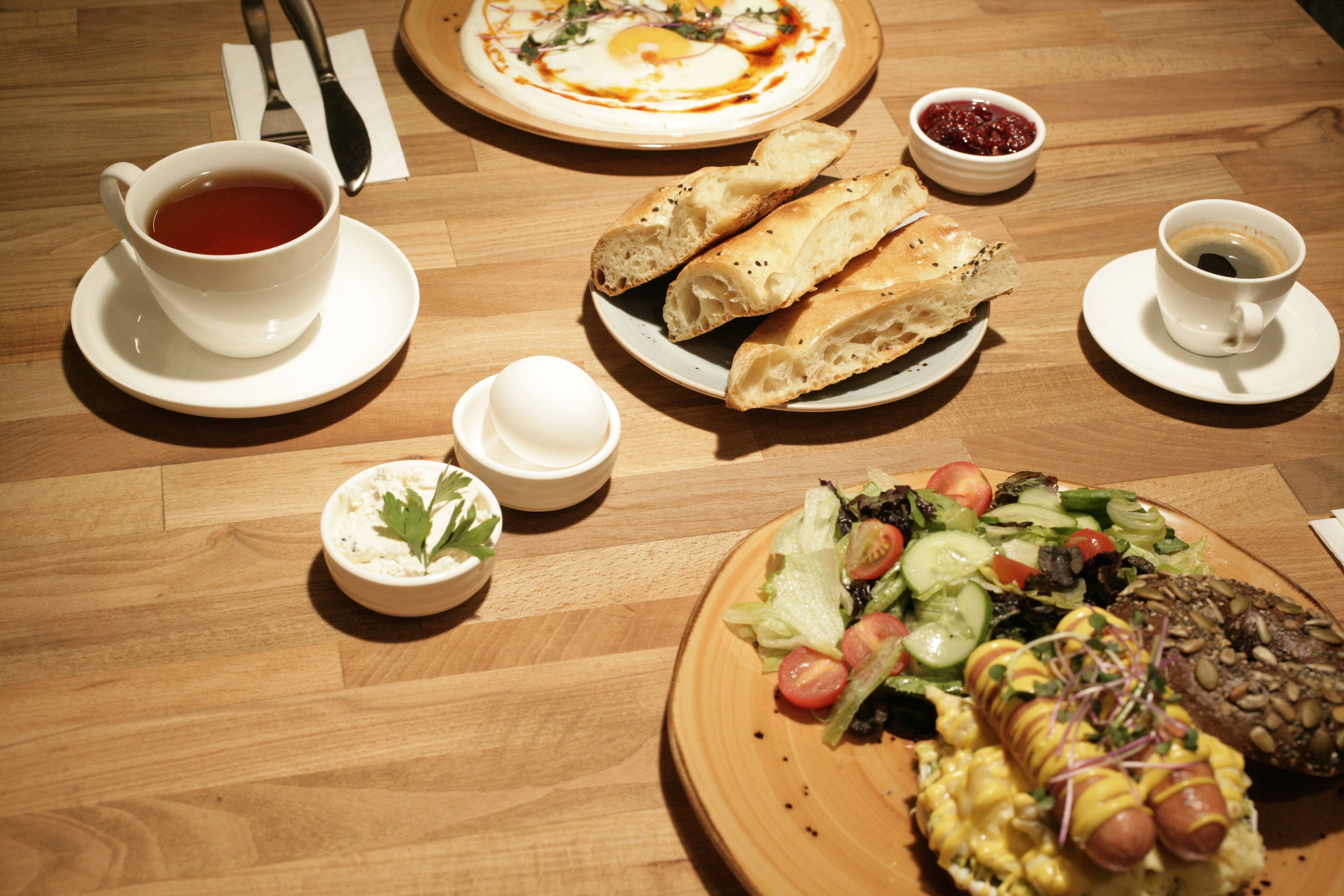 A wooden table with various breakfast items including bread, a hot dog, a salad, and two cups of coffee.