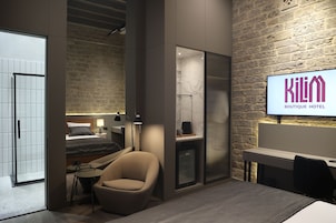 Een moderne hotelkamer met een bed, een zithoek met een stoel en tafel, een klein bureau en een groot televisiescherm waarop de naam van het hotel staat.