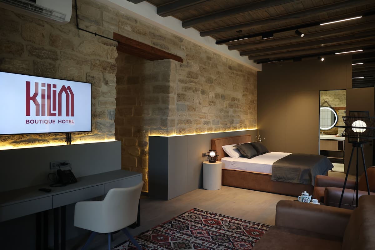 Ein modernes Hotelzimmer mit einer Steinwand, einem Bett mit Holz-Kopfseitenverkleidung, einem Schreibtisch mit Stuhl, einem Nachttisch mit Lampe und einem großen Spiegel.