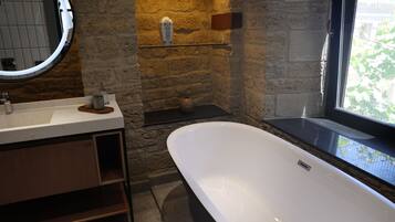 A bathroom with a white bathtub, a round mirror, a stone wall, and a window with a view of greenery.