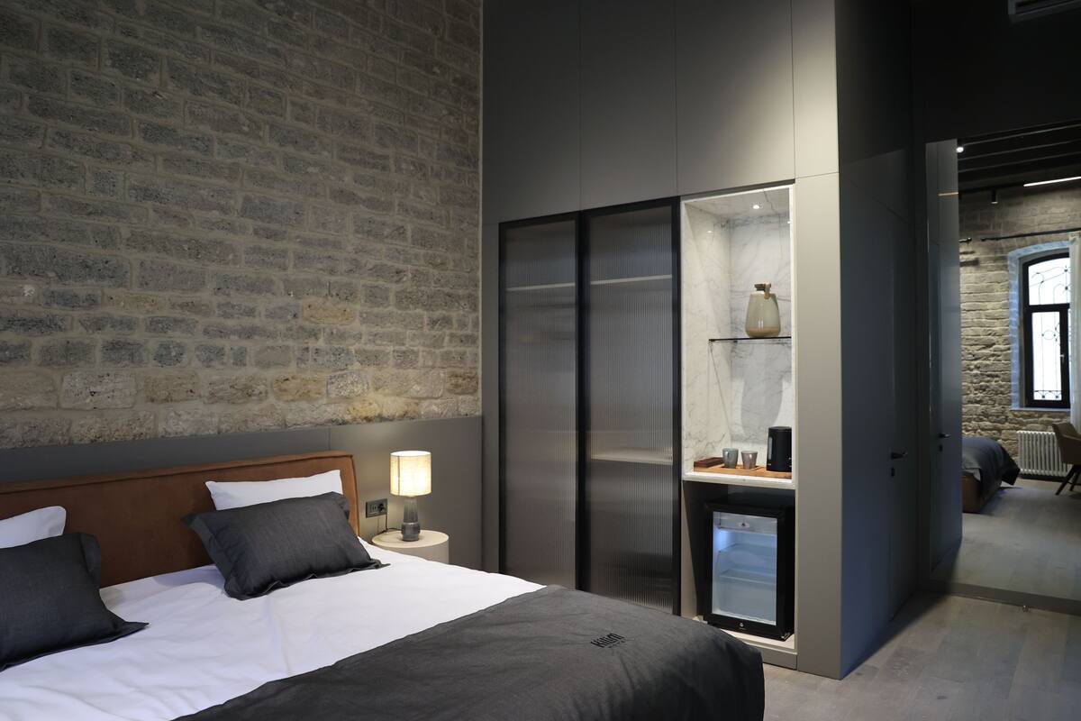 Ein modernes Hotelzimmer mit Backsteinwand, einem Bett mit weißer und dunkelgrauer Bettwäsche, einem Nachttisch mit Lampe und einem Schiebetürenschrank.