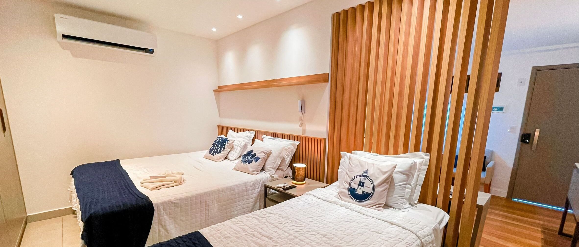 A hotel room with two beds, a wooden slat partition, an air conditioning unit, and a bedside table with a lamp.