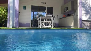 Une piscine avec une chaise blanche au centre, une porte donnant sur un espace abrité et un coin cuisine équipé de placards et d’appareils électroménagers.
