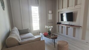 A living room with a sofa, a coffee table, a television, and a window with blinds. - Ecolodge Barbuda - Natural Pool & Beach (Saint-François)