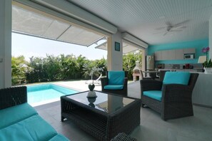 A modern kitchen with grey cabinets and a microwave, a dining area with wicker furniture and teal cushions, and a poolside seating area with wicker sofas and a glass coffee table. - Villa the Lodge - Lagon Saint-françois (Saint-François)