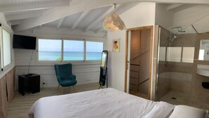 A bedroom with a bed, a chair, a TV, a window with a view of the ocean, and a shower area. - Villa Clara (Le Gosier)