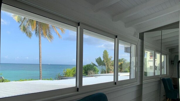 A room with large windows offering a view of the ocean and palm trees. - Villa Clara (Le Gosier)
