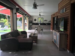 Ett rymligt vardagsrum med en soffa, ett matbord och en tv. Längre in i rummet finns ett kök med en klocka och ett fönster.