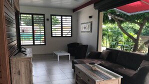 A living room with a sofa, coffee table, and a cabinet. There are two windows with blinds, one wall-mounted artwork, and a red umbrella outside. - Villa Ambre (Saint-François)