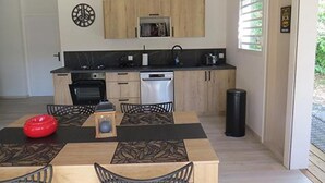 A modern kitchen with wooden cabinets, a built-in oven, and a dishwasher. There is a dining table with chairs and a red bowl on it. - Ecolodge Anguila - Natural Pool & Beach (Saint-François)