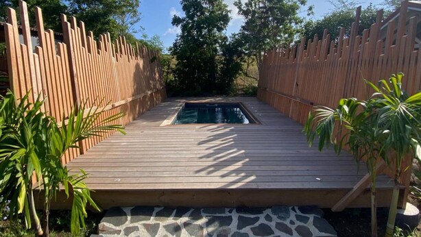 A wooden deck leading to a swimming pool with a stone border, surrounded by a wooden fence and greenery. - Ecolodge Anguila - Natural Pool & Beach (Saint-François)