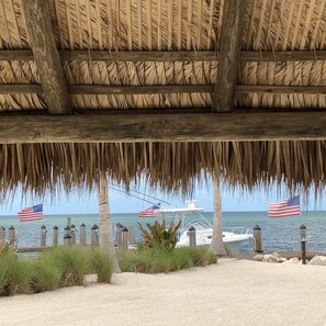 A beach scene with a thatched roof structure, a sandy beach, and a view of the ocean with a boat and American flags. - 2 Beautiful 4 Bed 3 Bath Pool Home w Free Dock (Marathon)