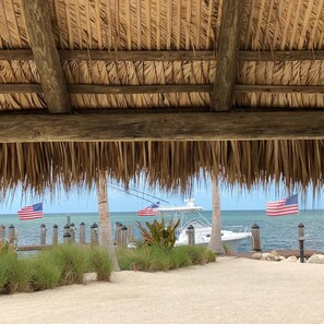 A beachside view with a thatched roof structure, a boat docked in the water, and American flags. - 3 Beautiful 4 Bed 3 Bath Pool Home w Free Dock (Marathon)