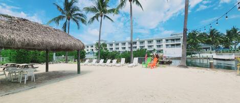 A beach area with lounge chairs, a thatched-roof structure, palm trees, and a hotel building in the background.