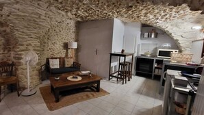 Una habitación rústica con paredes de piedra, un sofá, una mesa de madera, un ventilador y una zona de cocina con estufa y refrigerador.