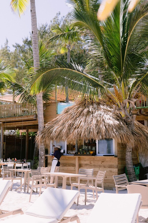 A beachside restaurant with outdoor seating, palm trees, and a thatched roof structure.
