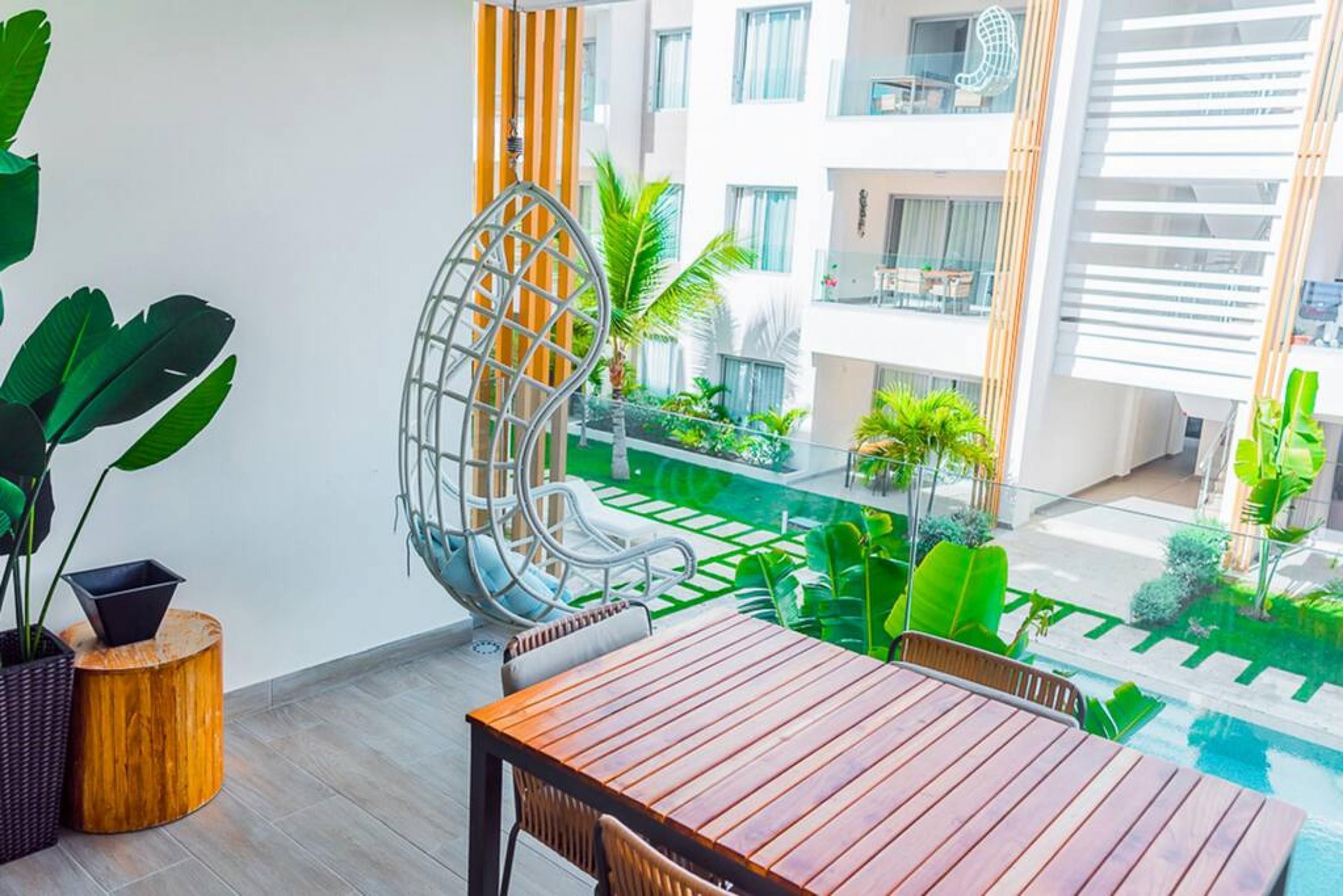 A modern outdoor dining area with a wooden table and chairs, surrounded by lush greenery and a view of a residential building.