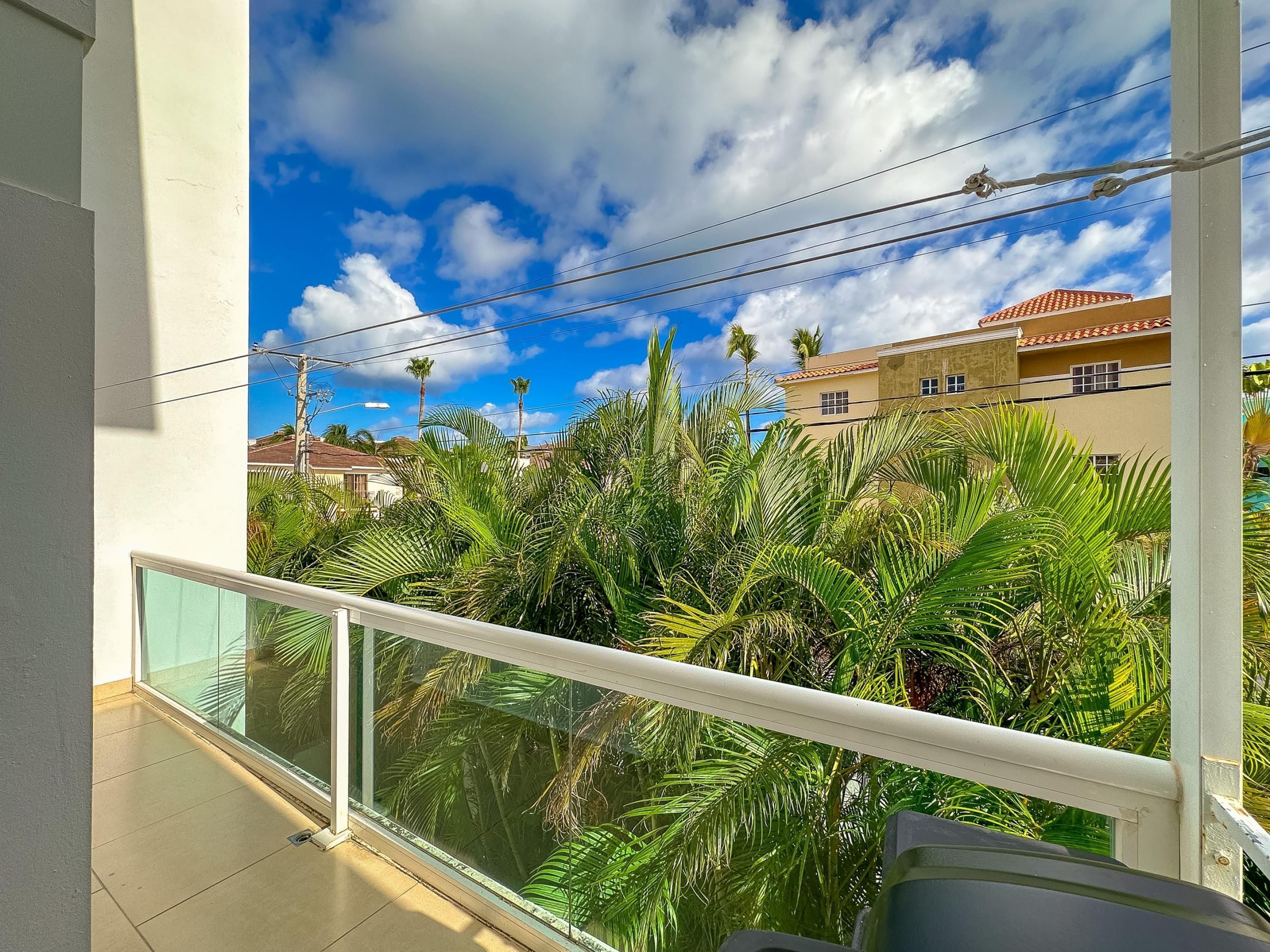 A balcony with a glass railing overlooking palm trees and a building with a red-tiled roof.